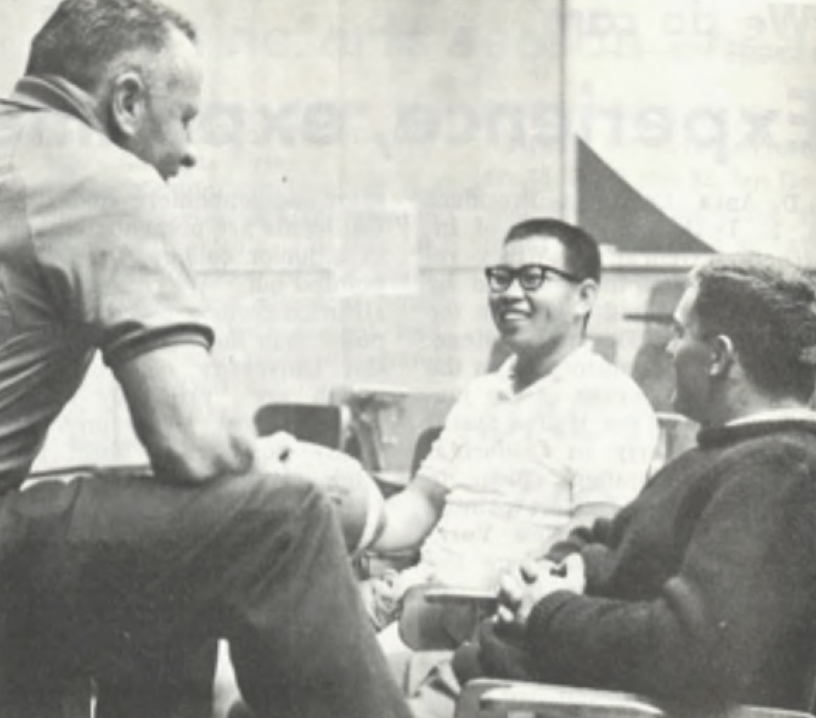 Three White and Asian men are sitting and engaged in conversation. The man on the left is sitting on a table or elevated surface, while the other two are seated in chairs. 