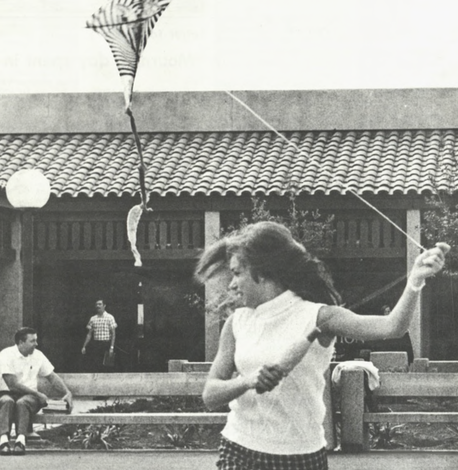 A young woman in a sleeveless top and plaid skirt flies a kite outside a tiled-roof building, with a few people sitting on a bench in the background. Her hair is blowing in the wind as she enjoys a sunny day.