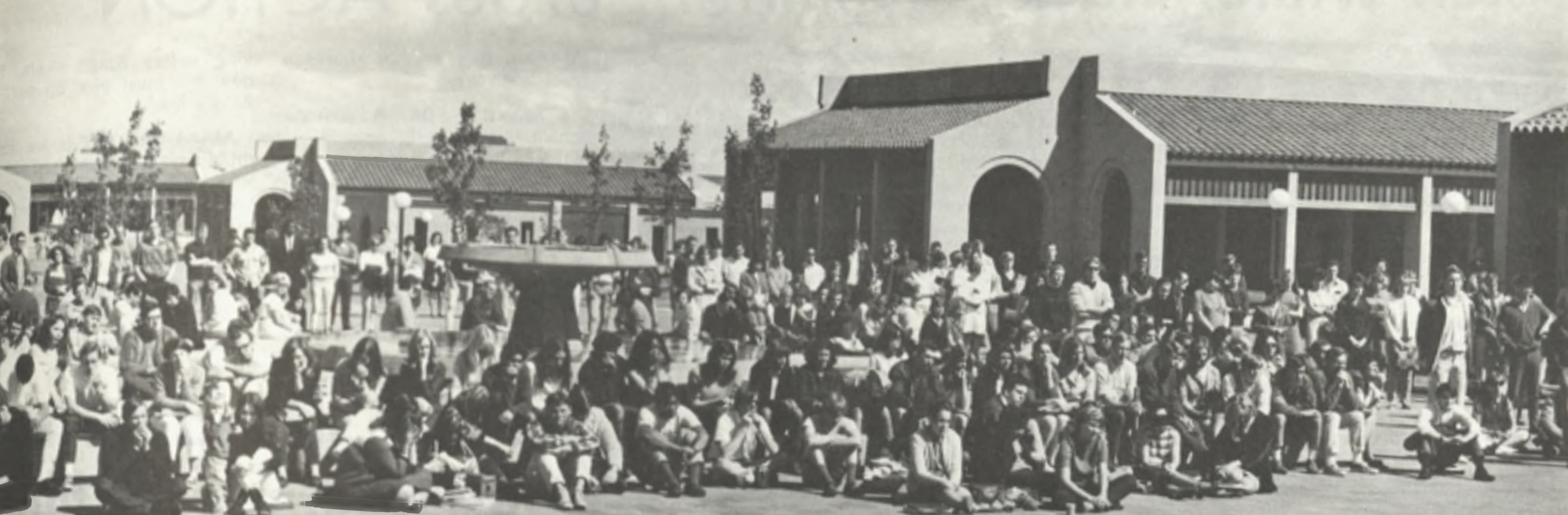 A large group of people, seated and standing, is gathered outdoors in front of a series of buildings with tiled roofs