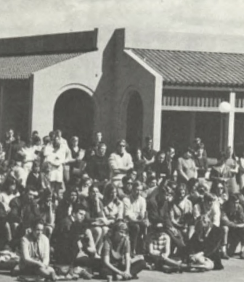 A large group of people, seated and standing, is gathered outdoors in front of a series of buildings with tiled roofs