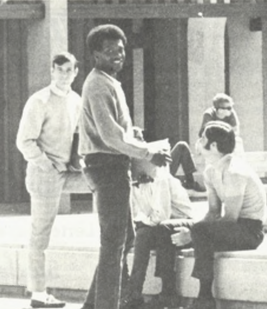 A group of students is gathered in an outdoor setting, casually interacting. One of them, a young Black man, is standing holding papers and smiling at the camera, while others are seated on stone benches.