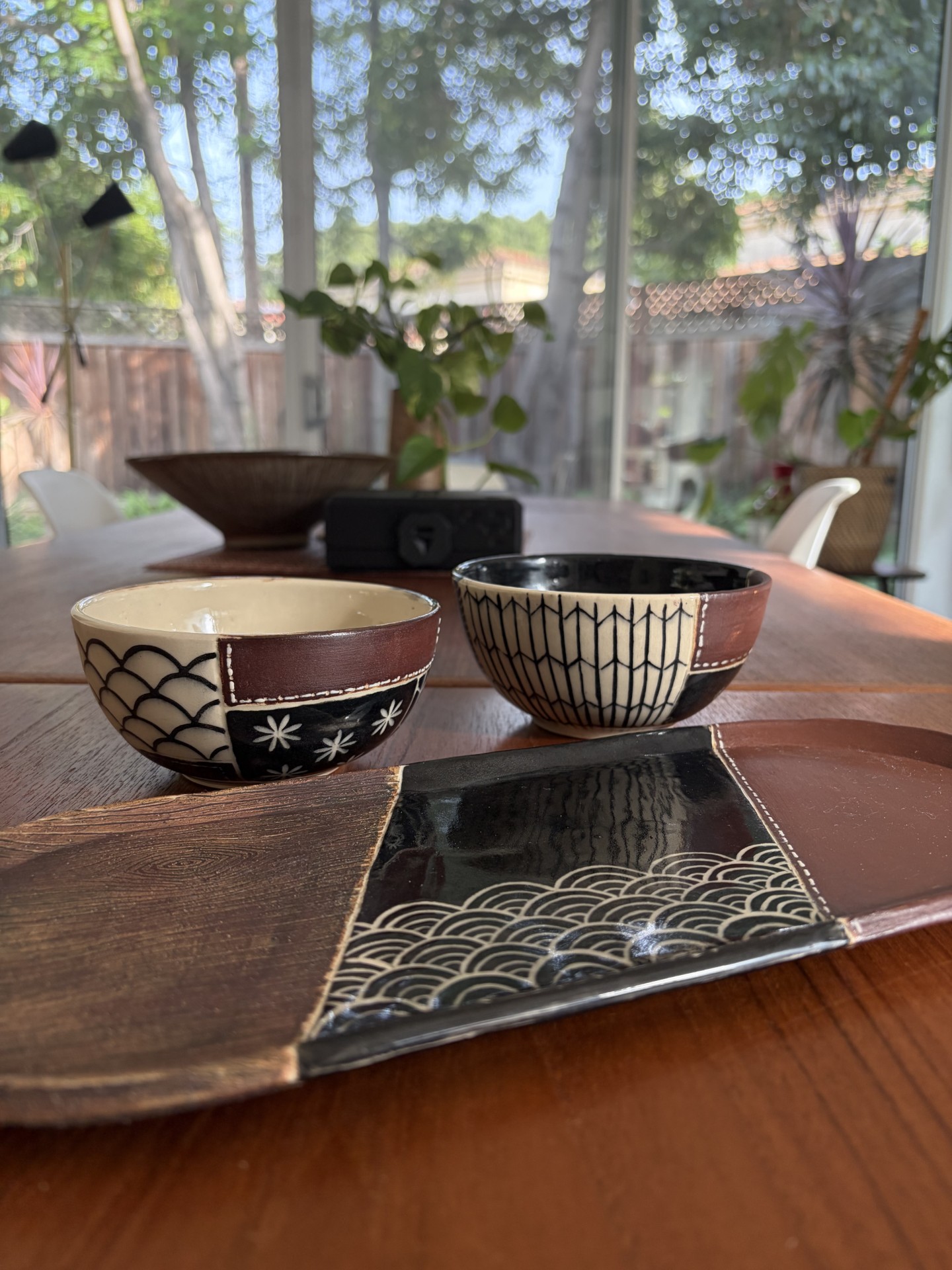 Two patterned ceramic bowls and a square tray with black and white geometric designs, displayed on a wooden table with a garden view behind