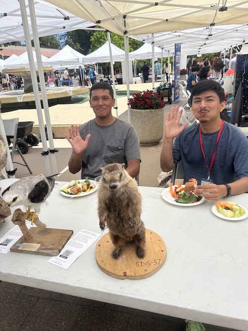 two young men waving at table with stuffed squirrel